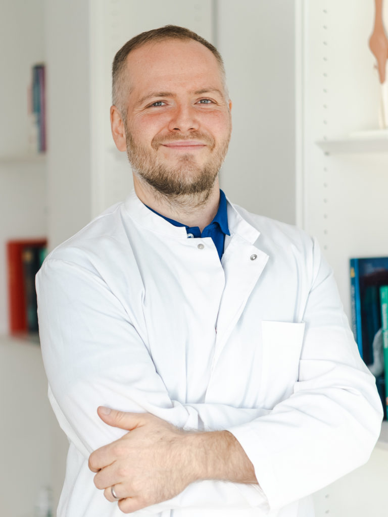 Dr. Michael Wunderlich, ein Orthopäde aus München in einem weißen Arztkittel, schaut in seiner Privatpraxis lächelnd in die Kamera.